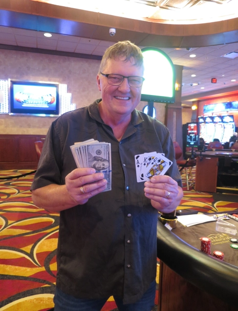 A smiling man stands in a casino holding a fan of cash in one hand and several playing cards, including an ace and a king, in the other hand. Casino tables and slot machines are visible in the background.