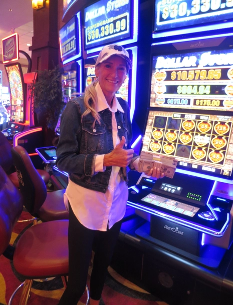 A smiling woman in a denim jacket and cap stands in front of a slot machine inside a casino, holding a stack of cash and giving a thumbs-up. Bright lights from the slot machines illuminate the background.