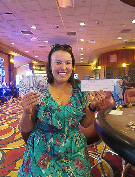 A smiling woman in a green patterned dress sits at a casino table, holding a fan of cash in one hand and a prize check in the other. The casino floor and slot machines are visible in the background.