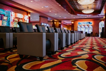 Rows of empty armchairs line a colorful, patterned carpet in a casino, with slot machines in the background and warm lighting overhead.