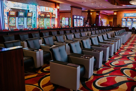 Rows of empty black and gray armchairs face slot machines in a brightly lit casino with colorful patterned carpet and illuminated gaming displays.
