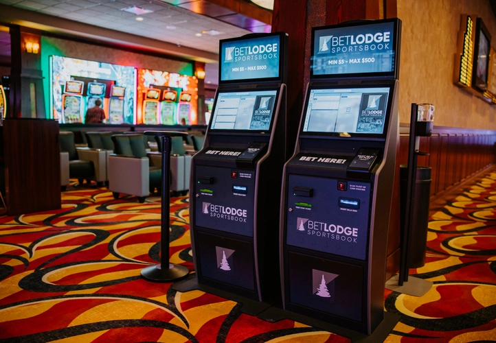 Two BetLodge Sportsbook betting kiosks in a casino, with colorful carpet and slot machines in the background. The kiosks display betting options and touchscreen interfaces.