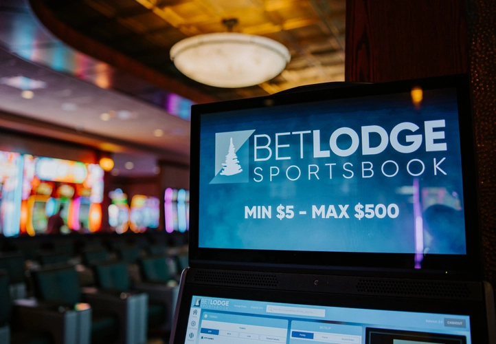 A sports betting kiosk screen displays "BETLODGE SPORTSBOOK, MIN $5 - MAX $500" in a dimly lit room with rows of empty seats and colorful slot machines in the background.