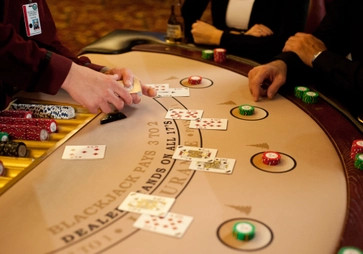 A casino dealer deals cards at a blackjack table, with several players' hands, poker chips, and drinks visible. The table surface displays game rules and payouts.
