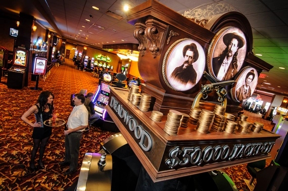 A casino floor with slot machines, a large decorative jackpot display showing $50,000 and old-time portraits, and two people talking near the display. The carpet has a red and orange pattern.