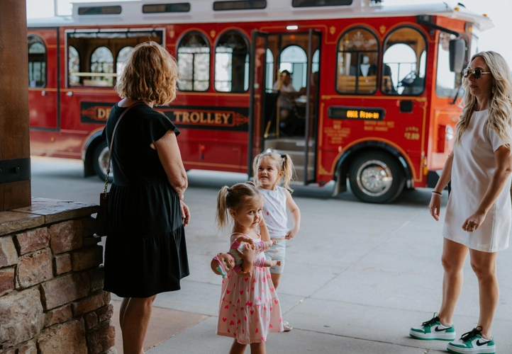 Two women and two young girls stand near a sidewalk, with a bright red trolley bus in the background. One woman wears a black dress, the other a white dress and sunglasses. The girls are in patterned dresses.
