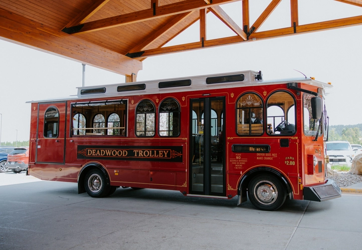 A vintage-style red trolley labeled "Deadwood Trolley" is parked under a wooden structure, ready for passengers. The trolley features gold detailing and large windows.
