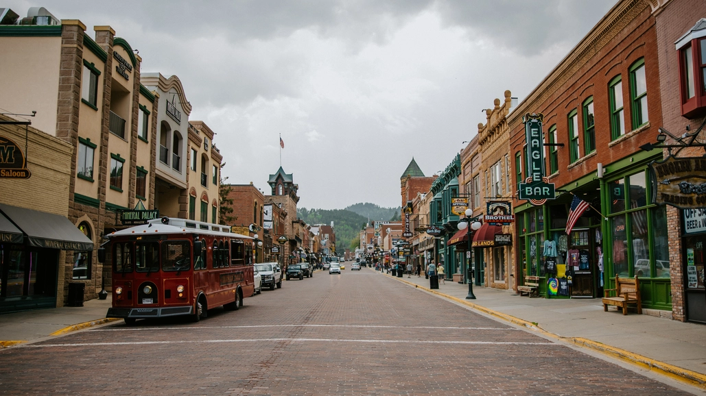 A brick-paved street lined with historic buildings, shops, and bars under a cloudy sky; a red trolley is parked on the left, and American flags are visible on some storefronts.
