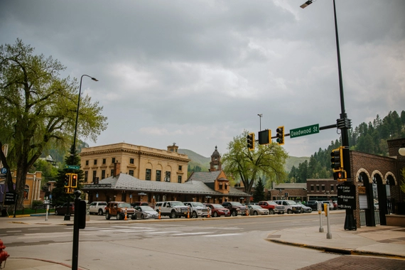 A street intersection with traffic lights in front of a historic-looking brick building. There are parked cars, green trees, and hills in the background under a cloudy sky. A street sign reads "Deadwood St.