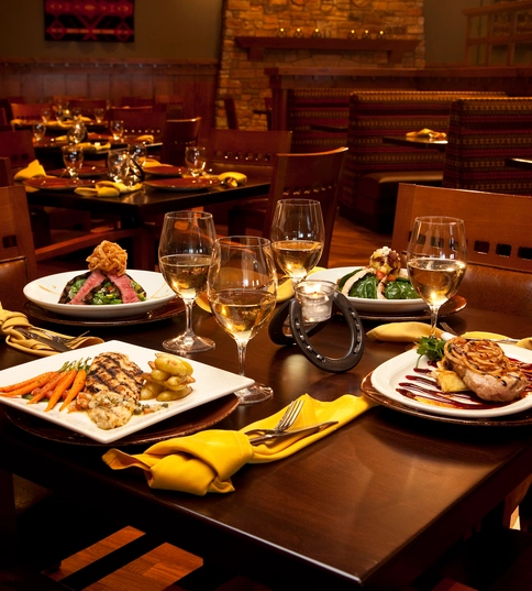 A restaurant table set with four gourmet meals, wine glasses, a bottle of white wine, yellow napkins, and elegant tableware. Warm lighting and wooden décor create a cozy, upscale dining atmosphere.