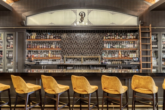 A modern bar with five tan leather stools, a black countertop, and shelves filled with various bottles of liquor. A rolling ladder leans against the shelves, and a patterned backsplash adds an elegant touch.