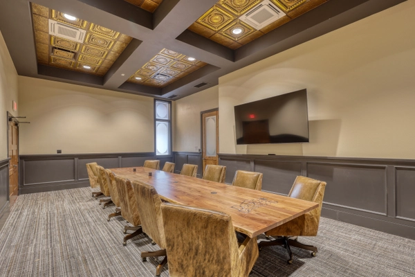 A modern conference room with a long wooden table, ten upholstered chairs, a large wall-mounted TV, decorative gold ceiling tiles, and a mix of natural and recessed lighting.