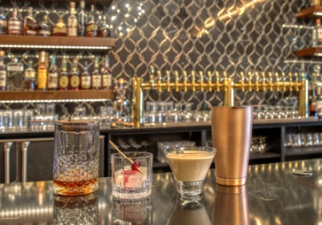 A bar countertop displays a glass with whiskey and ice, a glass with a maraschino cherry and large ice cube, a creamy cocktail in a short glass, and a copper shaker. Shelves of liquor bottles and glasses are in the background.