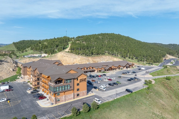A large, modern lodge-style hotel with a spacious parking lot, surrounded by green hills and trees under a clear blue sky. Several cars are parked in front of the hotel.