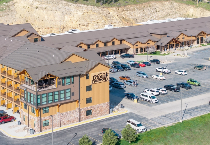 Aerial view of The Lodge at Deadwood, a large, multi-story hotel with a parking lot filled with cars, surrounded by hills and trees. The building has stone and wood accents, and balconies overlook the lot.