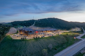 Aerial view of a large hotel and parking lot illuminated at dusk, surrounded by forested hills and located next to a road. Dozens of cars are parked outside, and the sky is tinged with evening light.