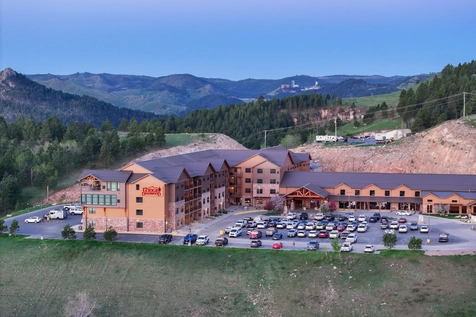 A large hotel with a parking lot full of cars sits among pine-covered hills and mountains under a clear blue sky. A few buildings and winding roads are visible in the background.