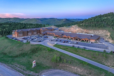 Aerial view of a large hotel and parking lot surrounded by green hills and forests, with a winding road in the foreground and mountains in the background under a clear sky at sunset.