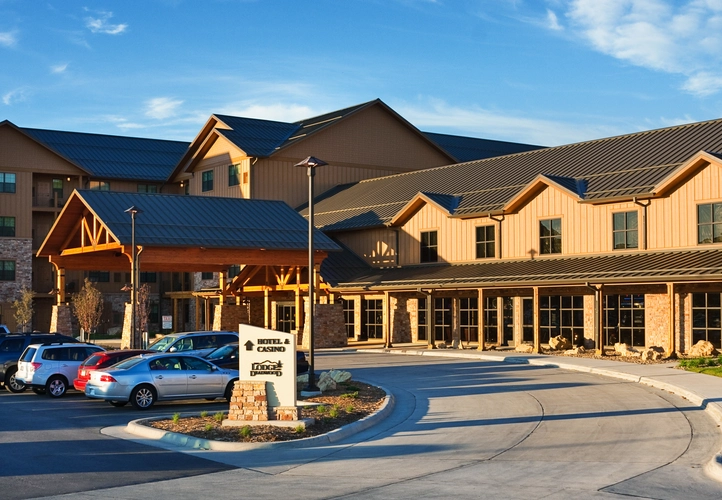 A modern tan and brown lodge-style building with large windows and a covered entrance. Several cars are parked out front, and a sign by the driveway reads “Hotel & Casino.” The scene is bright and sunny.