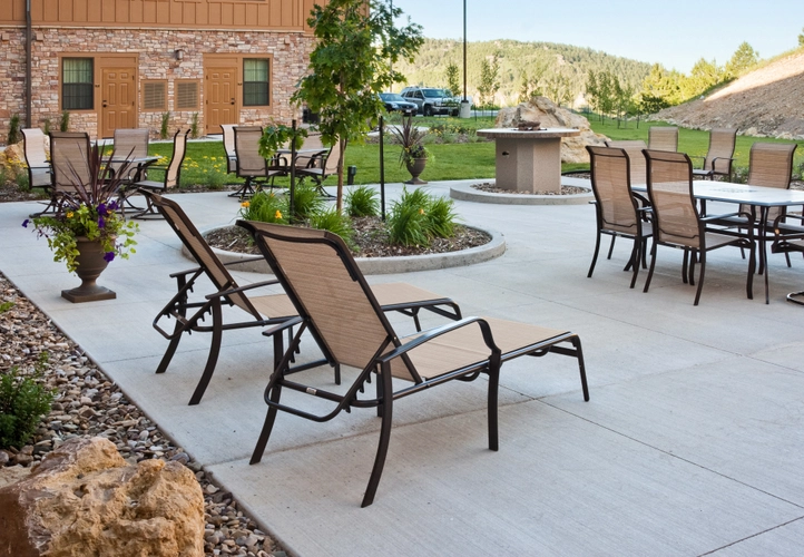 Outdoor patio with several tan lounge chairs and tables on a concrete surface, surrounded by landscaped plants and rocks, with a grassy area and hills in the background.