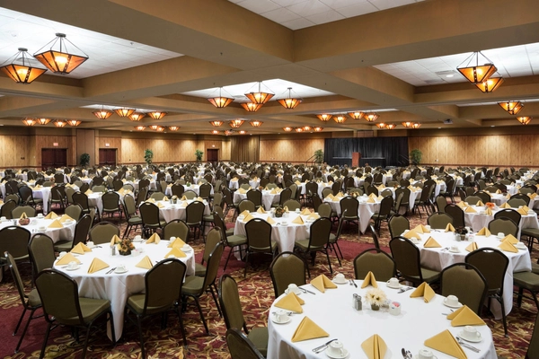A large banquet hall filled with many round tables covered in white tablecloths and set with yellow napkins, plates, cups, and silverware. The room is empty of people and lit by decorative ceiling lights.