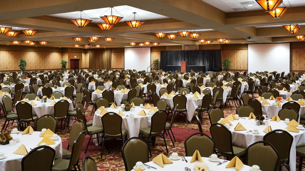 A large banquet hall with many round tables covered in white tablecloths, each set with yellow napkins, cups, and plates. Rows of chairs surround the tables, and warm ceiling lights illuminate the room.