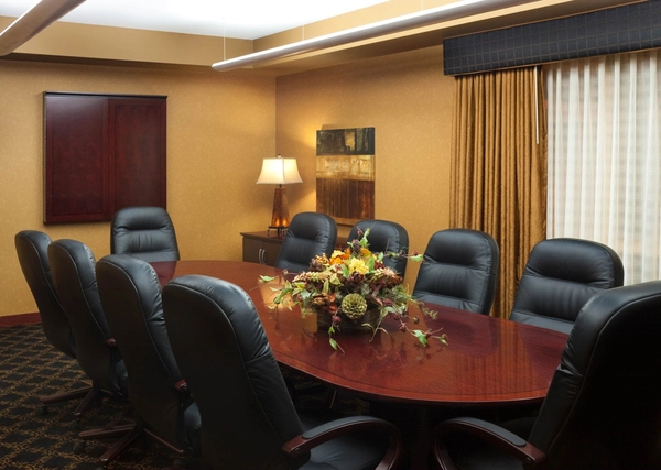 A conference room with a long wooden table, eight black leather chairs, a floral centerpiece, a lamp, abstract wall art, and gold curtains partially covering a window. The room has warm lighting and earth-toned walls.