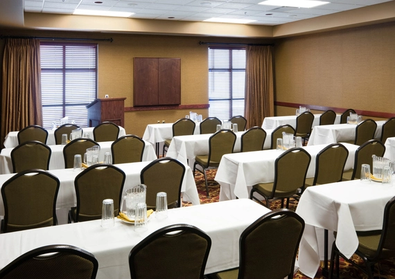 A conference room with rows of tables and chairs arranged classroom-style, each table covered with a white tablecloth, water pitchers, and glasses. Windows with closed blinds and a podium are at the front of the room.
