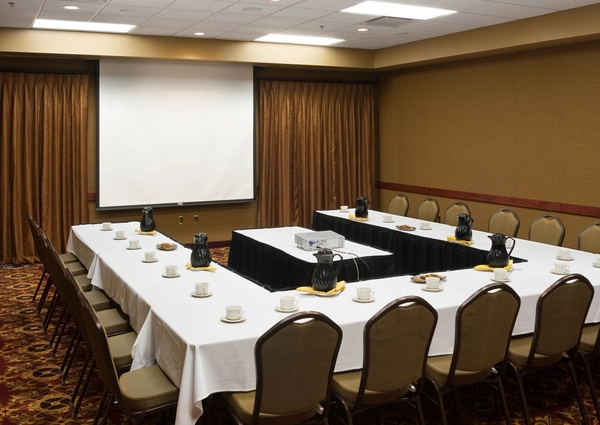 A conference room set up in a U-shape with tables covered in white cloths, coffee pitchers, cups, and plates, facing a projector and screen. Brown chairs and curtains complete the setting.