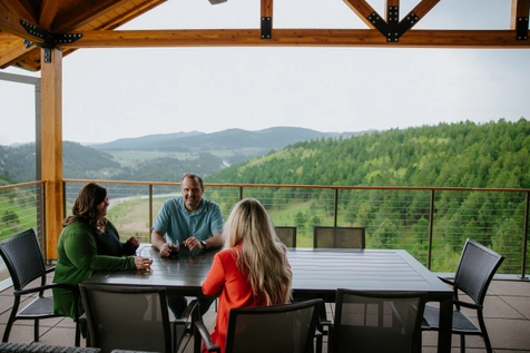 Three people sit around an outdoor table on a covered patio, talking and enjoying drinks, with a scenic view of green, forested hills and mountains in the background.