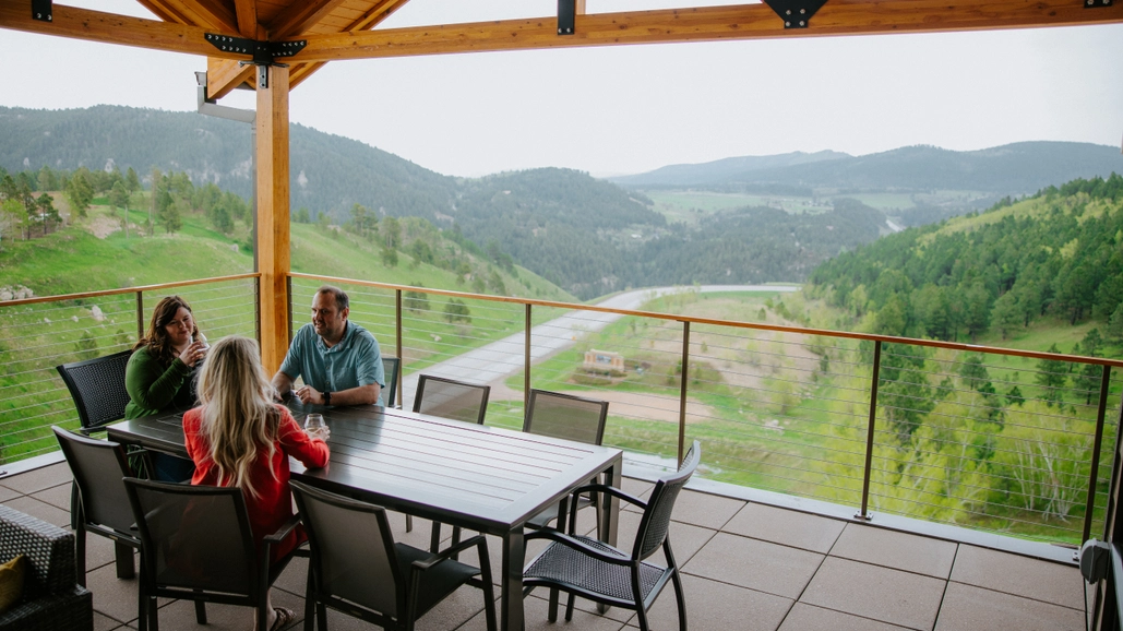 Three people sit around a patio table under a wooden pergola, talking and enjoying a scenic mountain and valley view with lush green hills in the background.