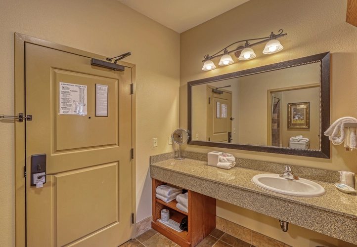 A hotel bathroom with a large mirror, granite countertop, sink, tissues, towels on open shelves below, a mounted towel ring, wall lights above the mirror, and a beige door with notices and a key lock.