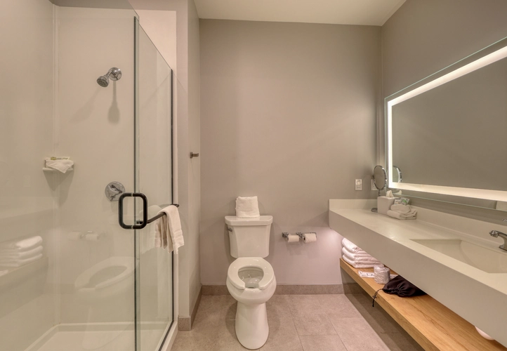 Modern bathroom with a glass-enclosed shower, toilet, large mirror with built-in lighting, white countertop with sink, wooden shelf below holding towels, and neutral beige walls and floors.