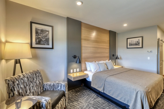 Modern hotel room with a neatly made bed, wood headboard, two nightstands, patterned armchair, floor lamp, and framed black-and-white photos on the walls. The lighting is warm and inviting.