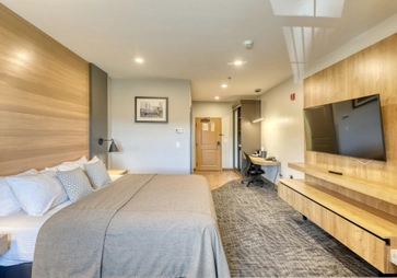 Modern hotel room with a king-sized bed, neutral bedding, a large wall-mounted TV, wooden furniture, a desk with a chair, and warmly lit decor. The door and a framed picture are visible in the background.