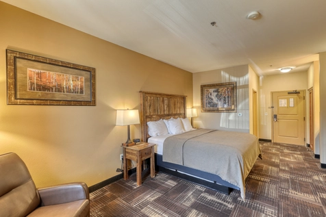 A warmly lit hotel room with a king-size bed, wooden headboard, two bedside tables with lamps, a cushioned chair, patterned carpet, and framed artwork on the walls near the entrance door.