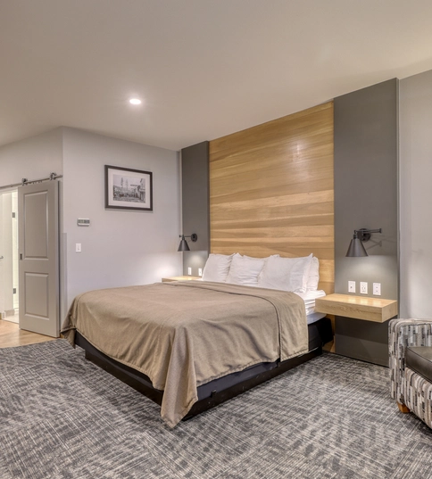 A modern hotel room with a large bed, beige bedding, a patterned armchair, wall-mounted lamps, framed black-and-white photos, wooden accent wall, and a door leading to a well-lit bathroom.