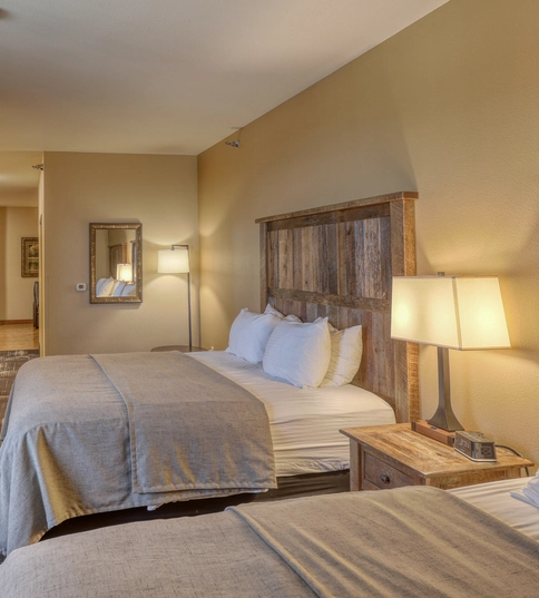 A hotel room with two double beds, wooden headboards, a nightstand with a lamp, a wall-mounted TV on a dresser, and neutral-colored walls and carpet. The entry door and a hallway are visible in the background.