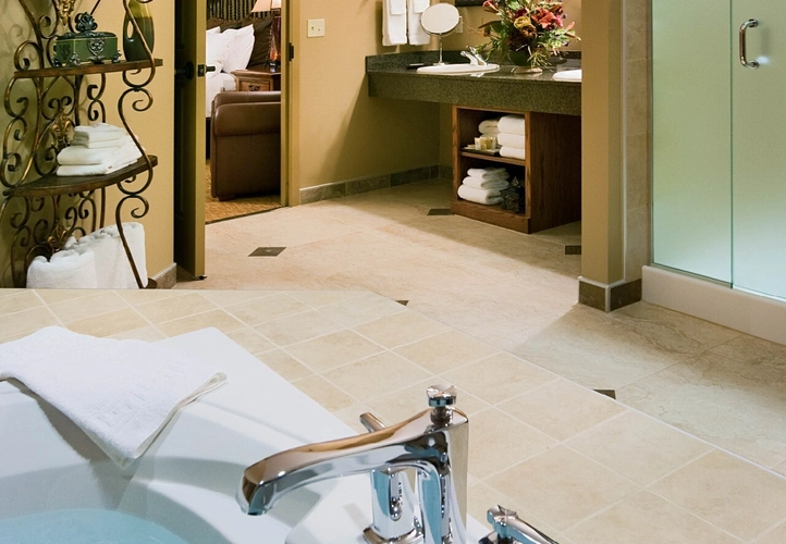 A spacious bathroom with a large bathtub filled with water, modern chrome fixtures, a glass shower, dual sinks with a large mirror, beige walls and floor tiles, and decorative shelves with plants and towels.
