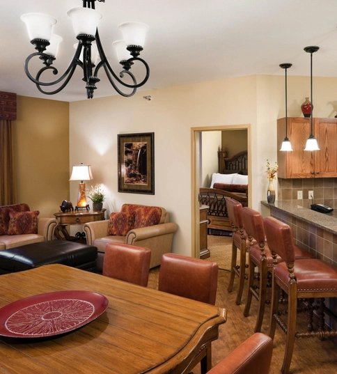 Open-concept living area with a dining table, kitchen with bar seating, and a living room. Brown leather chairs, wood cabinets, and modern lighting fixtures create a warm, cozy atmosphere. Bedroom visible in the background.