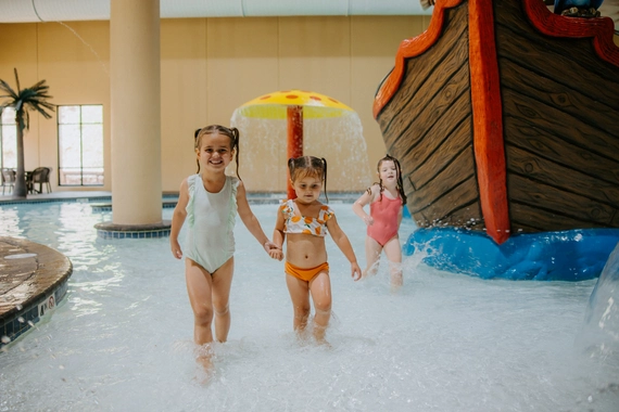 Three young girls in swimsuits play in shallow water at an indoor pool with a pirate ship and mushroom fountain in the background. Two girls hold hands and smile, while the third follows behind.