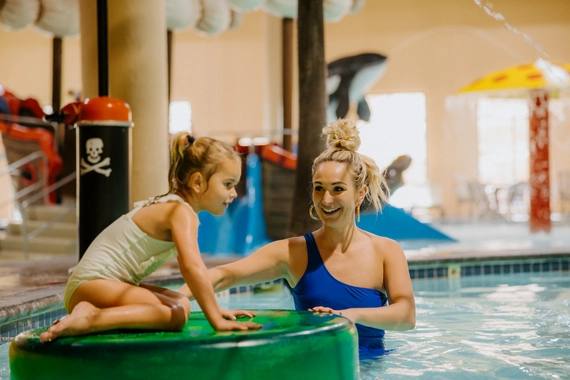 A woman in a blue swimsuit smiles at a young girl sitting on a green float in an indoor pool, with colorful play structures and water features in the background.