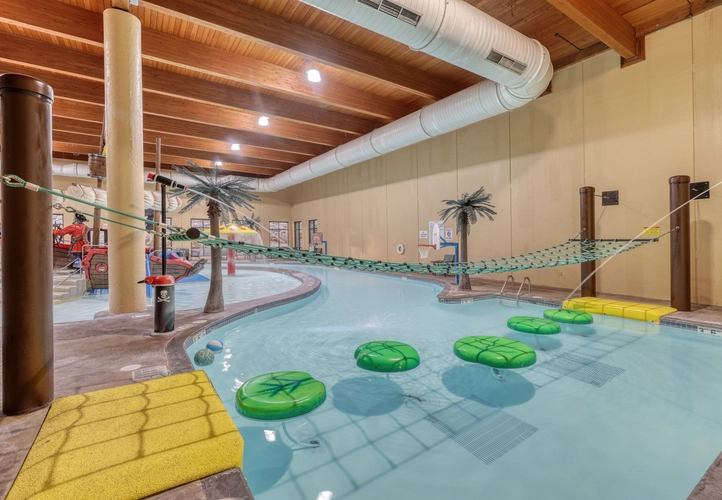 A pool obstacle course complete with floating lily pads and a rope net hanging above them. Water guns off to the side of the pool are spraying water on the course.
