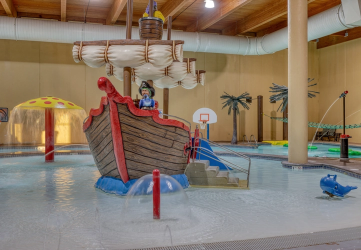 A fake pirate ship in the shallow end of the pool. There is a fake pirate at the front of the ship with a blue bird. A pool mushroom is off to the left of the ship and it's spraying water. There is a whale spring rider to the right of the ship.