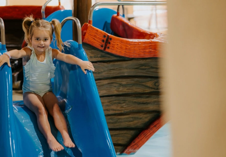 A little girl sitting at the top of a blue pool slide. She is wearing a light blue swimsuit and her hair is in pigtails.