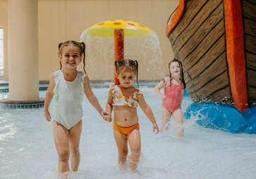 Two little girls are walking in the pool holding hands. Another one is walking behind them near a fake pirate ship.  A pool mushroom spraying out water is in the background.