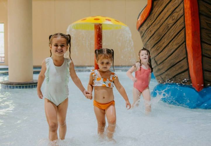 Two little girls are walking in the pool holding hands. Another one is walking behind them near a fake pirate ship.  A pool mushroom spraying out water is in the background.